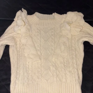 Vit stickad tröja med volangdetaljer - Superfin vit stickad tröja med grovt kabelstickat mönster och volanger på axlarna. Tröjan har rund halsringning och långa ärmar. Perfekt för dig som gillar en mysig och trendig look under kyliga dagar.