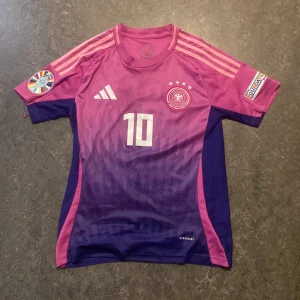 Tysklands Musiala #10 fotbollströja Adidas - Säljer en officiell Tyskland fotbollströja från Adidas med Musiala och nummer 10 på ryggen. Tröjan är i lila och rosa toner med vita detaljer, korta ärmar och snyggt geometriskt mönster framtill. Märkt med AEROREADY-teknologi och patchar på båda ärmarna.