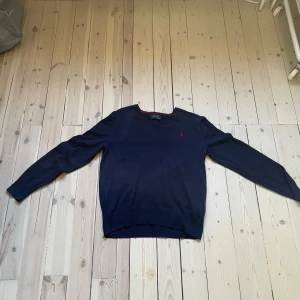 Mörkblå stickad tröja från Polo Ralph Lauren - Snygg mörkblå stickad tröja från Polo Ralph Lauren med klassisk v-ringning och röd broderad logga på bröstet. Tröjan har långa ärmar och ribbade muddar vid ärmslut och nederkant. Perfekt för lager-på-lager under hösten och vintern. Storlek L 14/16.