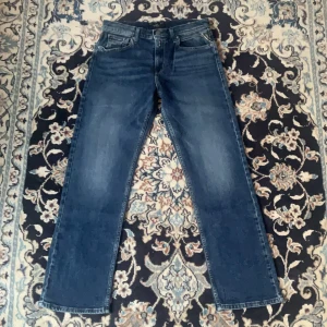 Blå raka jeans från Replay - Snygga blå jeans från Replay med klassisk femficksdesign och raka ben. Jeansen har en mellanblå tvätt med lätt slitning och tydliga sömmar. Märkespatch bak i midjan och normal passform. Perfekta för dig som gillar en tidlös jeanslook.