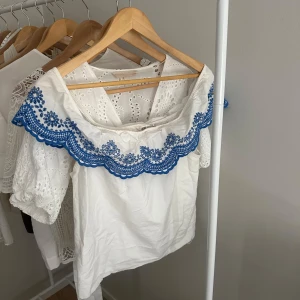 Vit offshoulder blus med blå brodyr - Superfin vit blus med offshoulder-modell och blå broderade blommor längs den vida kragen. Luftig och somrig känsla med korta ärmar och romantiska detaljer. Perfekt för dig som vill sticka ut med en söt och unik topp.