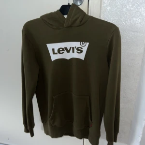 Olivgrön Levi's hoodie med logga - Snygg olivgrön hoodie från Levi's med stor vit logga på bröstet. Klassisk känguruficka framtill och huva. Mjuk insida och långärmad modell, perfekt för chill dagar. Passar dig som gillar streetwear och enkel stil.