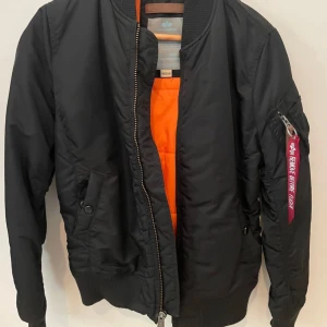 Svart bomberjacka från Alpha Industries - Säljer en svart bomberjacka från Alpha Industries med klassisk orange insida. Jackan har dragkedja, fickor med tryckknapp och ikonisk röd tag på ärmen. Perfekt för dig som vill ha en streetig och tidlös look.