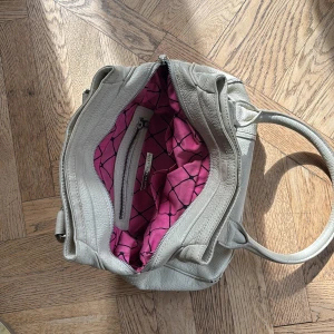 Beige skinnväska Marc by Marc Jacobs - Snygg beige handväska i äkta skinn från Marc by Marc Jacobs. Väskan har dubbla handtag, dragkedja framtill och coolt rosa innerfoder med svart grafiskt mönster. Perfekt storlek för allt du behöver ha med dig och riktigt stilren look.