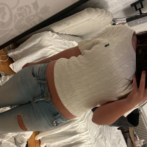 Vit kabelstickad tröja Polo Ralph Lauren - Vit kabelstickad tröja från Polo Ralph Lauren med kort ärm och klassisk rund hals. Tröjan har broderad marinblå logga på bröstet och ribbade muddar. Perfekt till jeans för en clean och fräsch look.