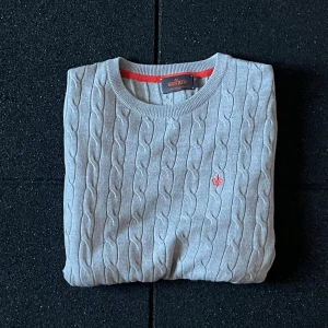 Grå kabelstickad tröja Morris M - Snygg grå kabelstickad tröja från Morris i 100% merinoull. Tröjan har rund hals, lång ärm och en röd broderad logga på bröstet. Perfekt för dig som gillar klassisk stil med en modern touch. Passar dig som vill ha något både bekvämt och stilrent.