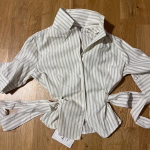 Randig figurformad skjorta med knyt i midjan - Snygg vit och beige randig figurformad skjorta från Fashion med knytband i midjan och klassisk krage. Skjortan har knappar framtill och trekvartsärmar. Perfekt för dig som vill ha en fräsch och trendig look med lite extra detalj i midjan. Går att knyta in vid midjan hur mycket som helst! Ger en snygg snatched look!! Helt ny med pris lapp!! PASSAR XS OCH S