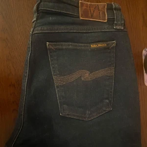 Mörkblå Nudie Jeans, W30 L32 - Säljer ett par mörkblå jeans från Nudie Jeans med klassisk bakficka och snygga kontrastsömmar. Modellen har dragkedjegylf och raka ben. Perfekta för dig som gillar stilrena och tidlösa jeans med schysst passform. Gylfen är sönder och behövs lagas
