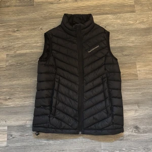 Svart dunväst från Peak Performance - Svart dunväst från Peak Performance med snyggt quiltat mönster och hög krage. Västen har två fickor med dragkedja och Peak Performance-logga på bröstet. Perfekt att slänga på över en hoodie när det är kyligt ute.