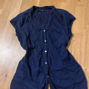 Mörkblå figurformad skjorta Monoprix strl 36 - Snygg mörkblå skjorta från Monoprix med korta ärmar och v-ringad hals. Framsidan har dekorativa veck och vita knappar hela vägen. Skjortan är gjord i mjuk bomull och har en avslappnad passform som är lätt att styla till jeans eller kjol.