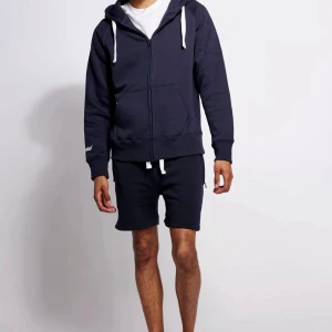 Mörkblå hoodie Russedress med dragkedja - Mörkblå hoodie från Russedress med vit snörning i huvan och broderad logga på ärmen. Tröjan har hel dragkedja, känguruficka och är gjord i mjuk bomullsblandning. Perfekt för chill dagar eller när du vill vara extra bekväm.