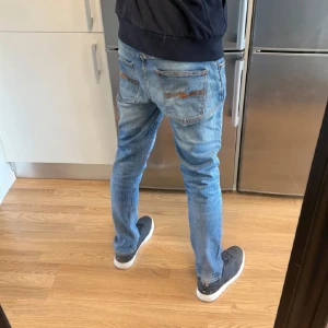 Nudie Jeans herr - Säljer ett par ljusblå jeans från Nudie Jeans med klassisk orange söm på bakfickorna och logga på baklappen. Jeansen har en rak passform och normal midja, tillverkade i mjukt bomullstyg. Perfekta för en avslappnad och stilren look.