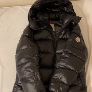 Svart Moncler Maya + NFC - Säljer en svart Moncler Maya jacka med glansig finish och huva. Jackan har dragkedja framtill, två sidofickor med dragkedja och klassisk Moncler-logga på ärmen. Fodret är dun och fjäder för extra värme. Perfekt för kalla dagar och riktigt snygg streetstil.