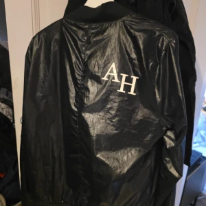 Adrian Hamond - Svart bomberjacka med emblem - Svart bomberjacka med glansig finish och ribbade muddar. På bröstet finns ett färgglatt emblem i rött, vitt och grönt, och på ryggen står stora vita bokstäver 'AH'. Jackan har dragkedja framtill och klassisk bomberkrage.