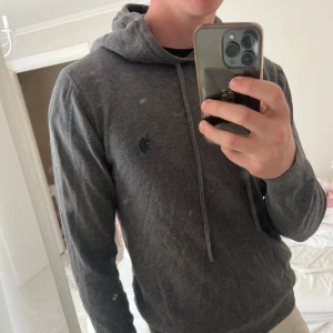 Stickad polo hoodie - Säljer en stilren grå hoodie från U.S. Polo Assn. med broderad logga på bröstet. Tröjan har huva med snörning, ribbade muddar och är tillverkad i mjuk bomull. Perfekt för dig som gillar en clean och klassisk look.