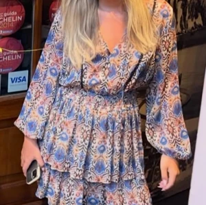 Mönstrad kortklänning med volanger - Superfin kortklänning med färgglatt mönster i blått, beige och rosa. Klänningen har v-ringning, långa vida ärmar och volangdetaljer i kjolen. Perfekt för dig som gillar boho-vibe och vill sticka ut lite extra. Använd en gång 