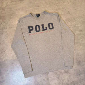 Grå långärmad tröja Polo Ralph Lauren - Snygg grå långärmad tröja från Polo Ralph Lauren med stor POLO-logga i mörkblått och röd text på bröstet. Tröjan har rund halsringning och är gjord i mjuk bomull, perfekt för en chill och stilren look.passar S men är strl 14-16.Pris kan diskuteras 