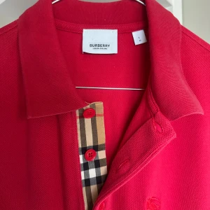 Röd pikétröja från Burberry, storlek L - Snygg röd pikétröja från Burberry med klassisk krage och korta ärmar. Tröjan har Burberrys ikoniska rutiga knappslå och röda knappar. Broderad logga på bröstet och skön bomullskvalitet. Perfekt för en clean och stilren look.