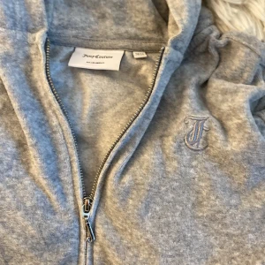 Grå zip hoodie från Juicy Couture och byxor  - Mysig grå hoodie från Juicy Couture med dragkedja och huva. Mjuk velourkänsla, broderad J på bröstet och praktiska fickor med knappar bak. Perfekt för chill dagar och streetstyle vibes. Byxorna följer med. Storlek 12-13 sitter som en 170-xs