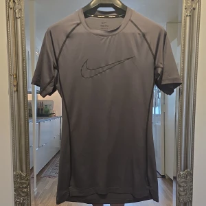 Nike Pro Dri-Fit grå träningstshirt - Nike Pro Dri-Fit t-shirt i mörkgrått med svart Nike-logga på bröstet. Tight passform och korta ärmar, perfekt för träning. Tillverkad i ett lätt och snabbtorkande syntetmaterial som andas. Snygga sömdetaljer och sportig look.