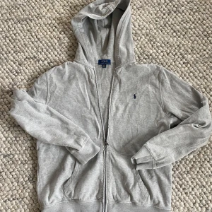 Grå hoodie från Polo Ralph Lauren  - Klassisk grå hoodie från Polo Ralph Lauren i storlek S/M. Tröjan har huva, dragkedja framtill och två fickor. Broderad marinblå logga på bröstet och mjukt bomullsmaterial. Perfekt för en chill och stilren look. Det står att den är XL i barnstorlek vilket korrelerar med ungefär S/M