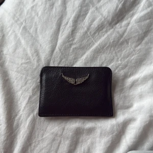 Svart kortfodral Zadig & Voltaire - Snyggt svart kortfodral i skinn från Zadig & Voltaire med silverfärgade vingar framtill. Stilren och smidig design med plats för flera kort. Använd och lite sliten men ingenting som syns. Äkta såklart 