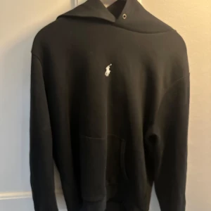 Svart hoodie från Ralph Lauren - Svart hoodie från Ralph Lauren med klassisk vit broderad logga på bröstet. Tröjan har huva, lång ärm och en stor magficka framtill. Perfekt för dig som gillar en enkel och stilren look.