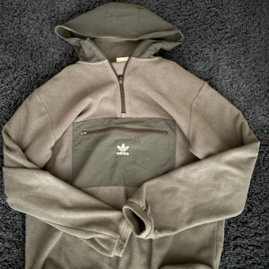 Grå fleecehoodie från Adidas - Säljer en grå fleecehoodie från Adidas med stor magficka och dragkedja framtill. Tröjan har huva, lång ärm och en clean Adidas-logga på fickan. Perfekt för kyliga dagar och riktigt skön att ha på sig.