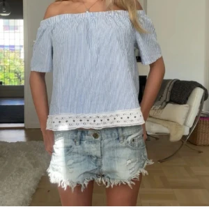 Randig offshoulder blus med spetskant - Superfin blus med blåvita ränder och offshoulder-modell. Toppen har korta ärmar och en bred spetskant längst ner som ger en söt touch. Perfekt till sommaren och enkel att matcha med jeansshorts eller kjol.