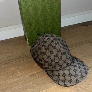Gucci keps  - Exklusiv keps från Gucci i beige och brunt med klassiskt GG-monogram över hela kepsen. Kepsen har gröna och röda ränder på sidan och är tillverkad i bomullsmix. Perfekt för dig som vill sticka ut med en ikonisk accessoar. Varan är så klart äkta! Finns lite defekter på kepsen priset är inte hugget i sten!