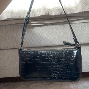 Svart axelväska i croco-imitation - Snygg svart axelväska med croco-mönstrad yta och justerbart axelband. Väskan har en stilren, avlång form och stängs med dragkedja upptill.