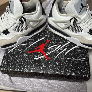 Nike Air Jordan 4 vit/svart/grå US10 - Nike Air Jordan 4 i vitt, svart och grått med klassisk meshpanel, svarta detaljer och Jumpman-logga på plösen. Skorna har snörning, platt sula och är tillverkade i skinn och syntet. Perfekt för dig som gillar streetwear och sneakers med ikonisk design.