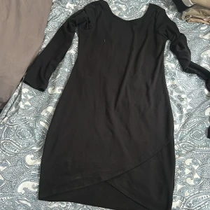 Svart långärmad omlottklänning - En svart, enkel och stilren klänning med lång ärm och omlott-detalj nertill. Klänningen har rundad halsringning och är figurnära i modellen. Den är något lägre i ryggen