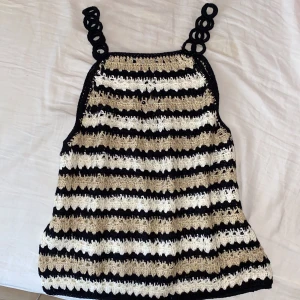 Randig virkad linnetopp - Säljer en unik virkad linnetopp med svarta, beige och vita ränder. Toppen har coola virkade axelband med ringdetaljer och är perfekt för varma dagar. Den är ärmlös och har en lätt A-linjeformad passning. Superfin till jeans eller kjol!
