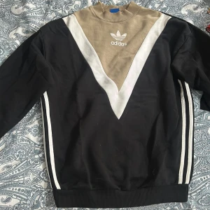Adidas svart och beige sweatshirt - Snygg Adidas sweatshirt i svart, beige och vitt med klassisk trefoil-logga på bröstet. Tröjan har en cool V-design framtill, ribbad krage och muddar samt vita tre streck längs sidorna. 