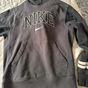 Svart hoodie från Nike XS - Svart hoodie från Nike i storlek XS med stor broderad logga och swoosh på bröstet. Klassisk känguruficka framtill och ribbade muddar. Ärmarna har vita ränder vid mudden. Perfekt för en chill och sportig look.
