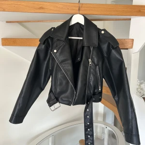 Svart skinnjacka med bälte och dragkedja - Snygg svart skinnjacka i klassisk bikerstil med silvriga dragkedjor, bred krage och axelklaffar. Jackan har ett justerbart bälte i midjan och coola detaljer som ger en edgy vibe. Perfekt för dig som vill ha en tidlös och trendig look.