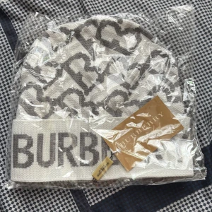 Vit och grå Burberry mössa - Snygg vit och grå mössa från Burberry med stort logotyp-tryck och mönster av stiliserade bokstäver. Mössan är stickad och har en bred uppvikt kant. Perfekt accessoar för att lyfta din vinterstil.