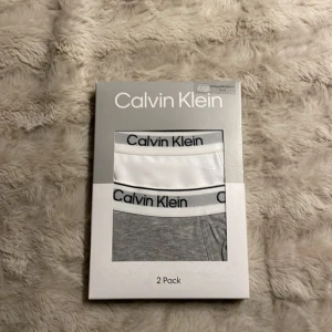 Calvin Klein 2-pack mjuka byxor - Tvåpack byxor från Calvin Klein i klassisk stil. Ena paret är helvita och det andra är grå, båda med bred resår i midjan med Calvin Klein-logga. Perfekta för chill eller träning, tillverkade i mjuk bomull för maximal komfort. Helt oanvänd!!