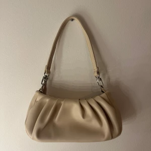 Beige axelväska i skinnimitation - Snygg beige axelväska med mjuk, rynkad design och silvriga metalldetaljer. Väskan har ett avtagbart axelband och är tillverkad i slät skinnimitation. Perfekt storlek för det viktigaste och ger en clean look till din outfit.