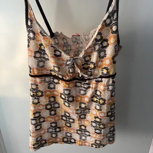 Mönstrad linnetopp med smala axelband - Säljer en cool linnetopp med smala svarta axelband och blommigt mönster i svart, gul och orange. Toppen har en lätt rynkad detalj vid bysten och svart band under bysten för extra form. Perfekt för varma dagar och sommarvibbar.