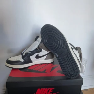Nike Air Jordan 1 High Mocha - Nike Air Jordan 1 High Mocha med svart, vit och brun färgkombination. Klassisk high-top siluett med svart swoosh, perforerad tåbox och snörning. Ovandel i skinn och mocka för en lyxig känsla. Kommer med extra vita skosnören. Perfekt för dig som gillar streetwear och sneakerskultur.