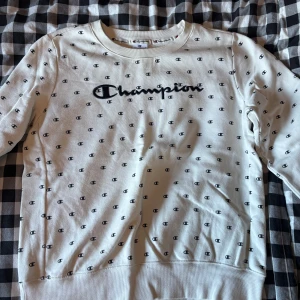 Vit sweatshirt från Champion XL - Vit sweatshirt från Champion i storlek XL med svart logotyptryck över bröstet och små svarta Champion-loggor över hela tröjan. Klassisk rund halsringning och långa ärmar. Mjuk insida och ribbade muddar vid ärmslut och nederkant. Perfekt för en chill och sportig look.