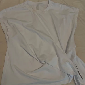 Vit ärmlös topp med knyt - Snygg vit topp med rund hals och ärmlös design. Toppen har en cool knytdetalj i sidan som ger en trendig twist. Perfekt att styla med jeans eller kjol för en clean look.