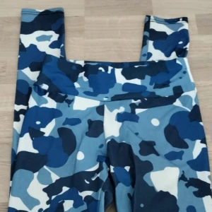 Blå camo leggings med hög midja - Snygga leggings med blått camouflage-mönster i olika nyanser av blått och vitt. De har hög midja och är tillverkade i ett stretchigt syntetmaterial som sitter tight och bekvämt. Perfekta för träning.