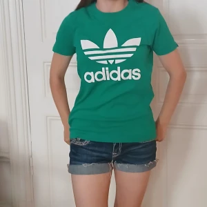 Grön Adidas t-shirt - Snygg grön t-shirt från Adidas med stor vit trefoil-logga och text framtill. Klassisk rund hals och korta ärmar. Skön passform och tillverkad i mjuk bomull, perfekt till jeans eller shorts för en sportig vibe. Köpt på en vintage butik förra året men har bara använt ett fåtal gånger, den är i bra skick men det vita tycket har några få "sprikor" men inget man tänker på❤️