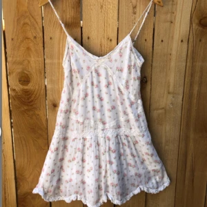 Vit blommig kortklänning med spets - Supersöt vit kortklänning med tunna justerbara axelband och små rosa blommor. Klänningen har spetsdetaljer längs kanterna och en volangig nederdel.  Vintage från Ralph Lauren!! Verkligen den perfekta sommarklänningen. Längd ca 65cm utan axelband