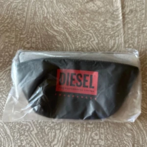 Svart magväska från Diesel - Cool svart magväska från Diesel fragrance med röd logga framtill. Väskan har justerbart band och dragkedja upptill.  Tillverkad i syntetmaterial med stilren look.