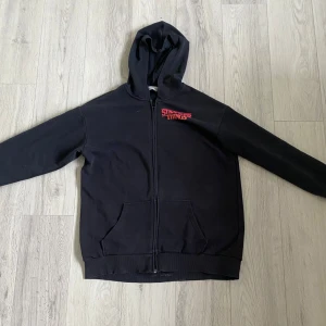 Svart Stranger Things hoodie från H&M - Svart hoodie från H&M med Stranger Things-tryck fram och stort färgglatt motiv på ryggen. Tröjan har huva, dragkedja och känguruficka. Perfekt för dig som älskar serien och vill ha en chill och snygg look.