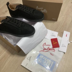 Svarta sneakers från Christian Louboutin - Snygga svarta sneakers från Christian Louboutin i mocka med klassisk röd sula och diskret logga. Skorna har rund tå, svarta skosnören och en stilren design som sticker ut med den ikoniska röda undersidan. Perfekta för dig som vill ha något exklusivt och trendigt.
